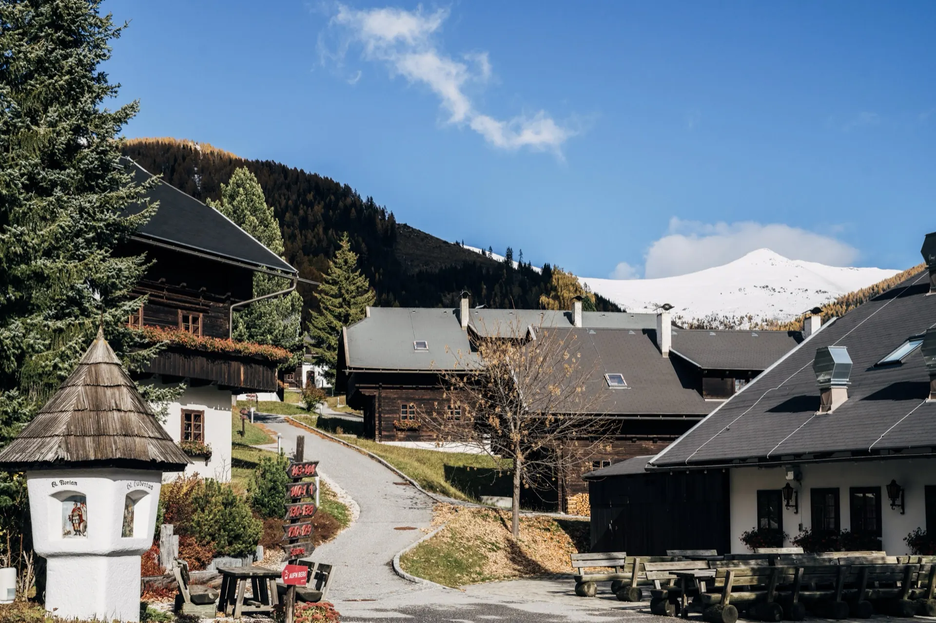 Dorfweg mit Bildstock und Holzhäusern im Herbst, verschneiter Berg dahinter
