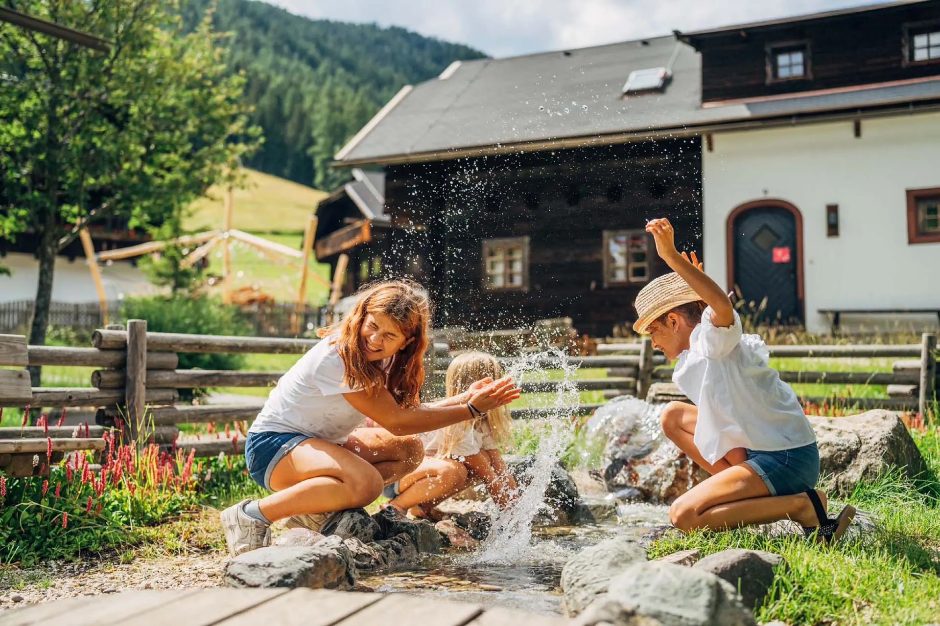 Mutter und Kinder spritzen mit Wasser am Bachlauf vor dem Holzhaus