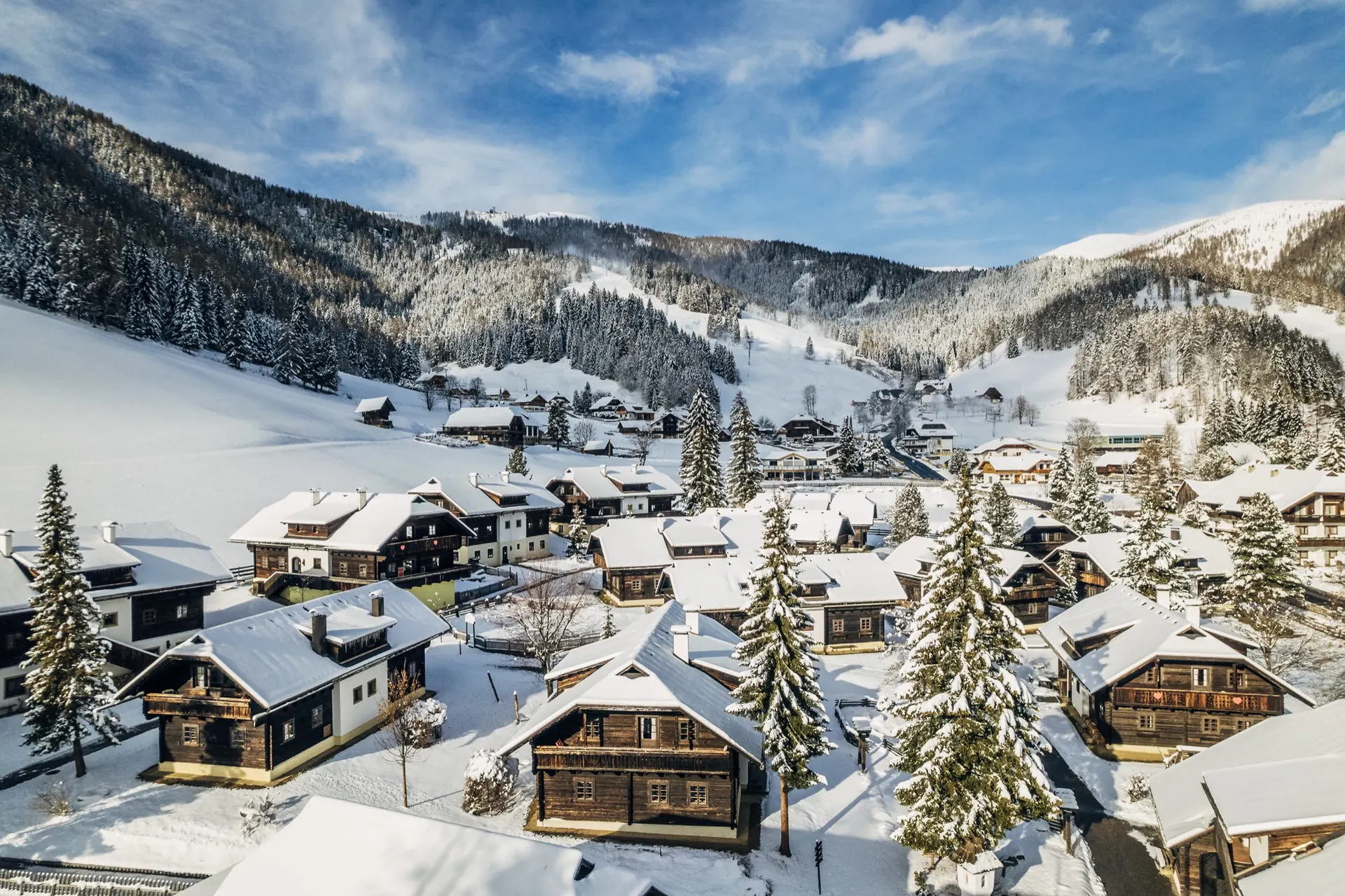 Kirchleitn aus der Luft im Winter: verschneite Holzhäuser, dahinter Tal und Nockberge