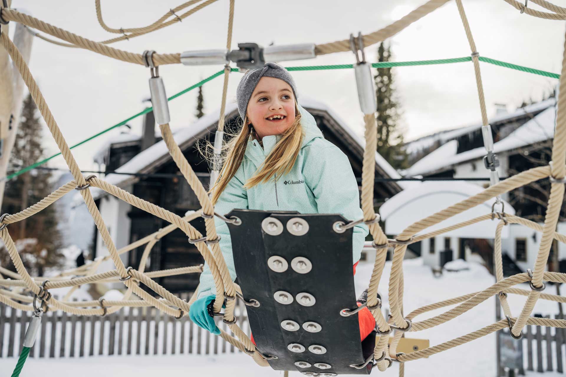 Mädchen am Kletterturm vor den verschneiten Ferienhäusern im Slow Travel Resort Kirchleitn in Bad Kleinkirchheim.